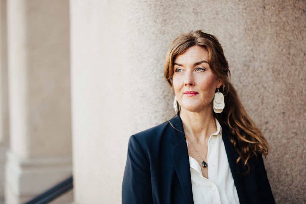 Sofia Djerf, legitimerad psykolog och handledare i Göteborg, lutar mot en ljus stenpelare och blickar snett framåt med ett eftertänksamt uttryck. Iklädd mörk kavaj och vit skjorta.